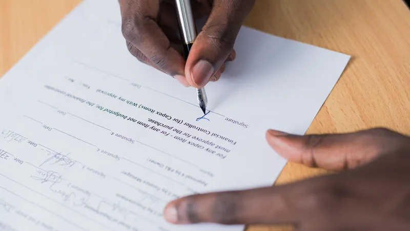 Close-up of two hands signing a printed document on a wooden table, with a pen.