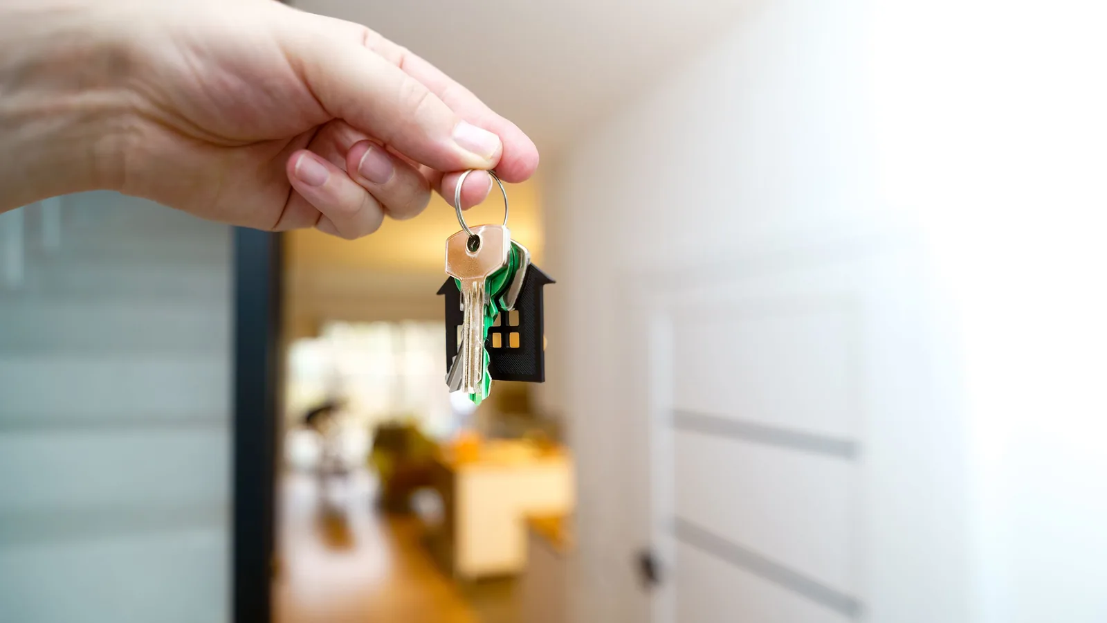 Une main tenant un porte-clés en forme de maison avec des clés, dans un couloir intérieur flou.