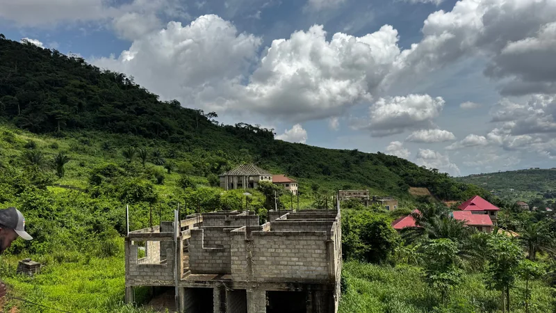 Construction inachevée en béton dans un paysage vallonné verdoyant avec végétation tropicale et toits rouges au loin.