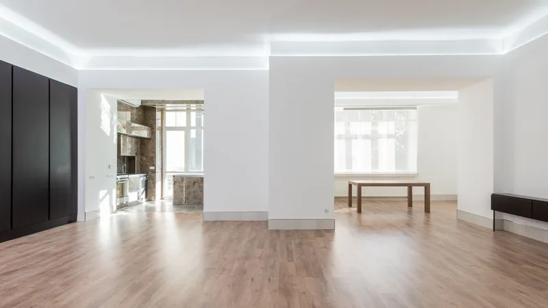 Grand salon vide au sol en parquet clair, murs blancs et plafonds à spots, sans aucun mobilier.
