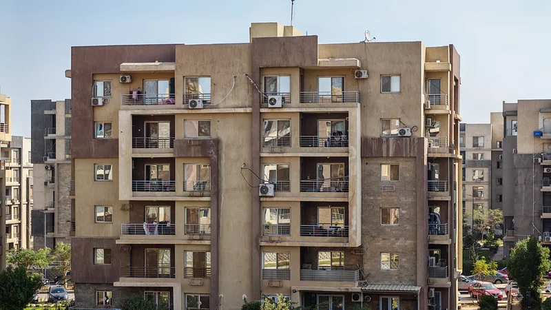 Multi-storey residential blocks with beige facades, balconies and air conditioners, greenery at street level, a street in the distance.
