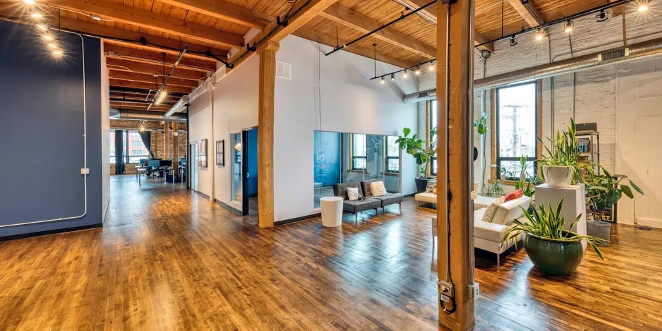 Spacious loft-style interior with wooden floors, exposed beams, and greenery, featuring seating areas and large windows.
