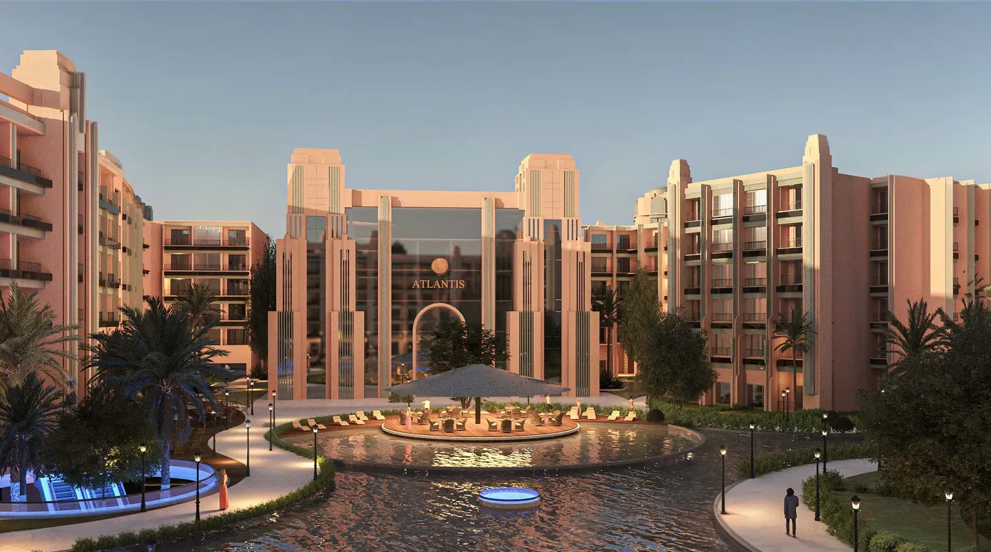 Modern luxury resort entrance with pinkish buildings, a water feature, palm trees, and outdoor seating under a large umbrella.