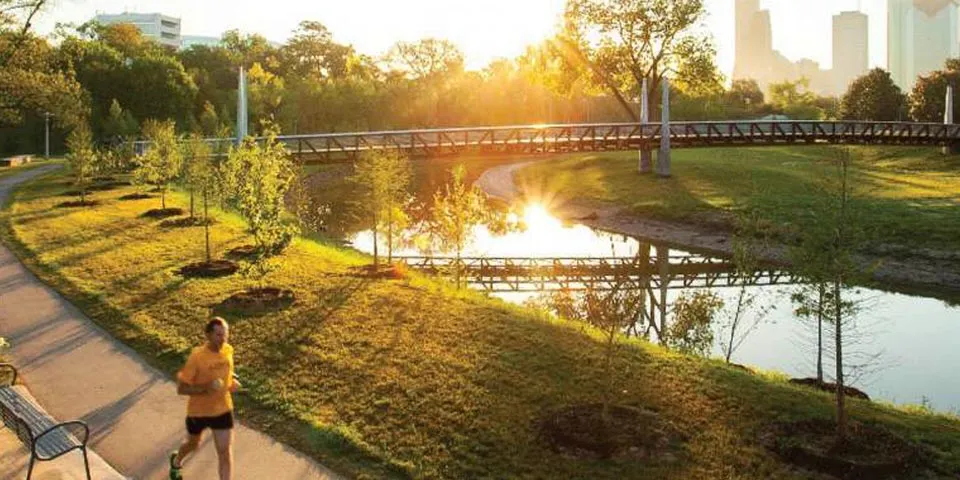 Runner jogging along a path by a tranquil river at sunrise, with city skyline in the background.