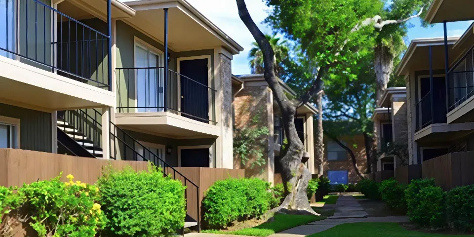 Two-story apartment buildings with balconies, surrounded by lush greenery and a paved walkway.