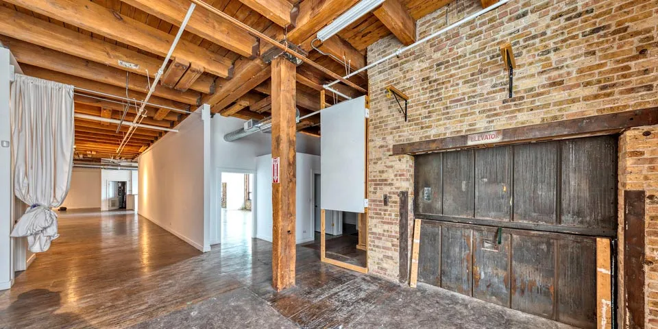 Spacious industrial loft with exposed brick walls, wooden beams, and large openings leading to adjacent rooms.