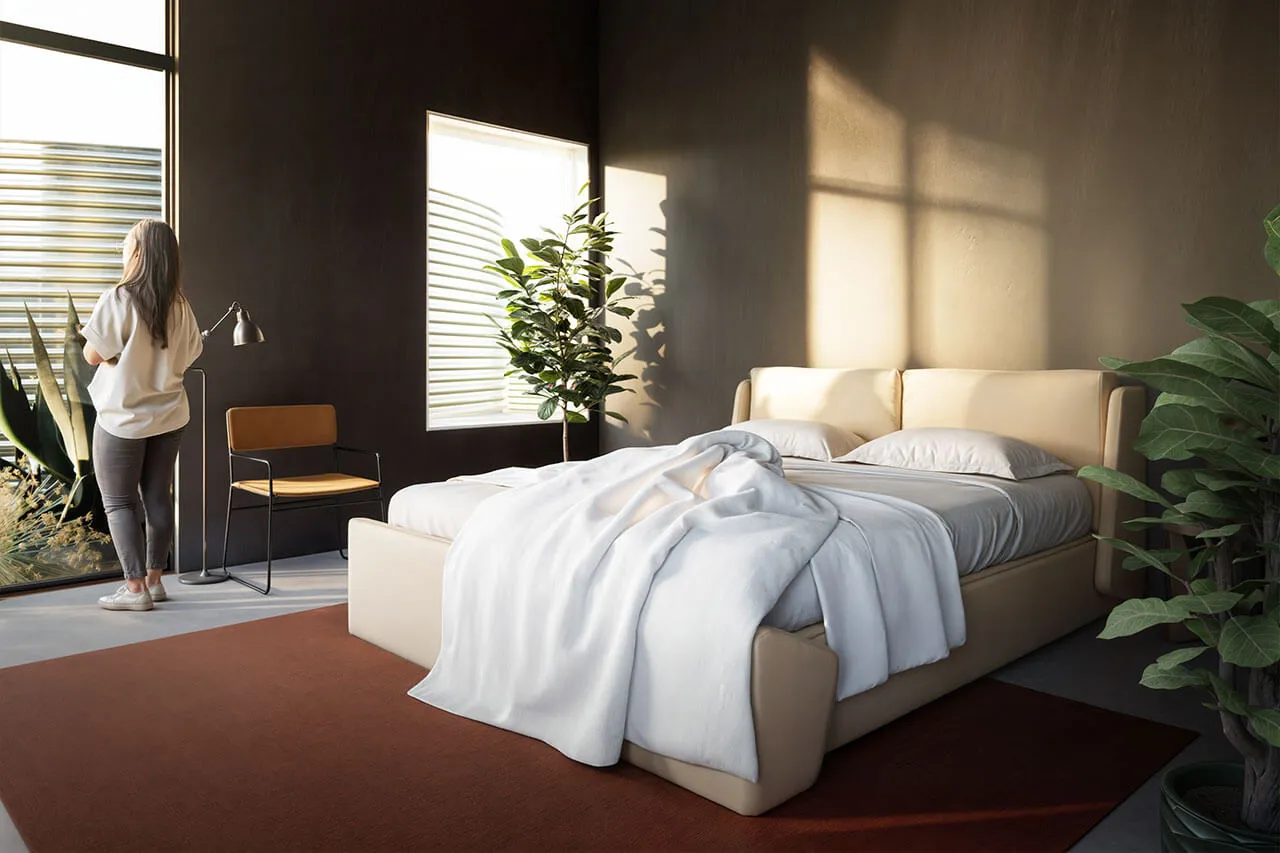 Woman with back to viewer looks out window in a bedroom with a beige bed, dark walls, and sunlight.