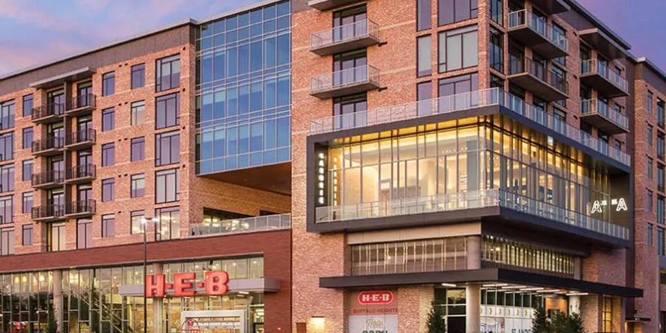 Modern brick building featuring H-E-B supermarket and residential apartments above, with large glass windows.